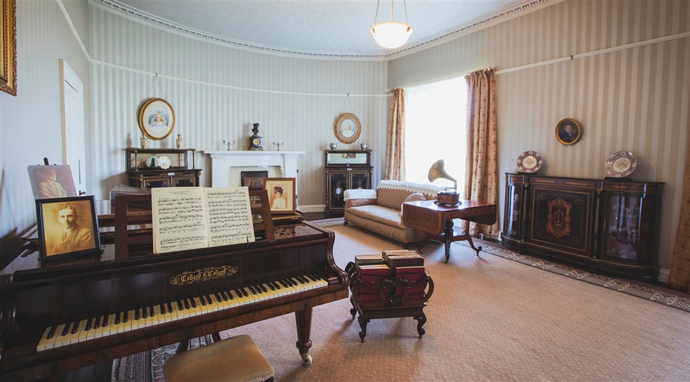 A front room with a piano, fireplace, gramophone and long pink curtains against stripey wallpaper at Abermydyr in Aberaeron, Ceredigion