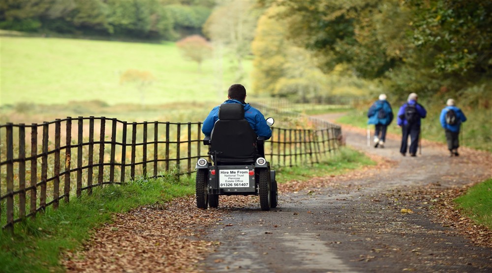 Someone riding a mobility scooter on a tarmacked path, in an autumnal woodland. at Bar Lodge in Helston, Cornwall