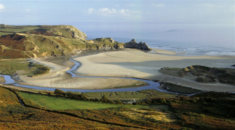 A view towards Three Cliffs Bay from near Penmaen, with a wide sandy bay visible between headlands and the ocean in the distance on a sunny day at Burrows Cottage in Swansea, South Wales