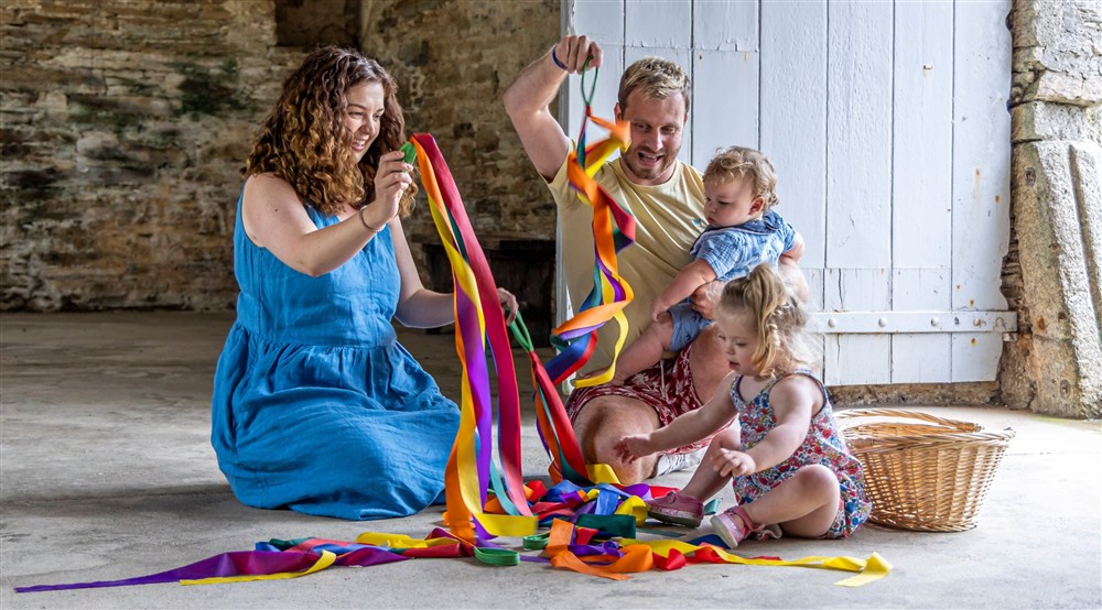 A family sit on a stone floor beside an open wooden door, playing with colourful streamers. The mother wears a long blue dress and is holding rainbow ribbons up for a toddler who is reaching for them. A baby boy is sitting behind on the fathers lap. at Ci