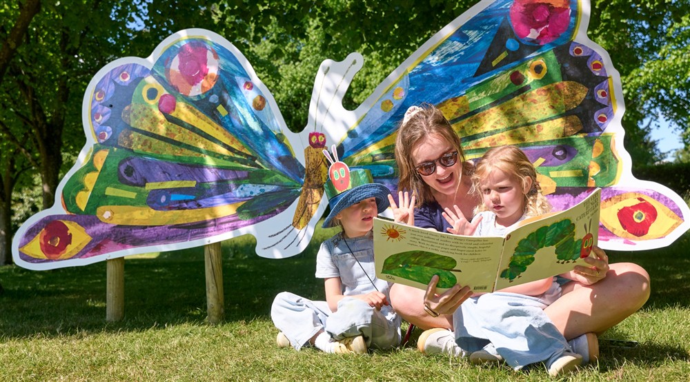 family sitting on the grass reading the Very Hungry Caterpillar book in front of a giant butterfly board at Cliveden New Cottage in Nr Maidenhead, Berkshire