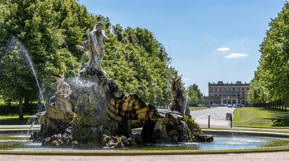 View of a large sculptural fountain in the foreground with green trees and a country house in the background with blue sky at Cliveden New Cottage in Nr Maidenhead, Berkshire