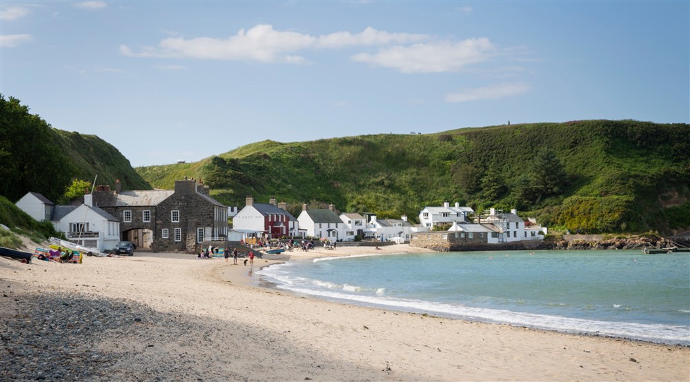 A view of the beach and coastal hamlet of Porthdinllaen with the coast visible on the right, a green cliffside behind the hamlet and people visible walking along the beach at Daron in Pwllheli, Gwynedd