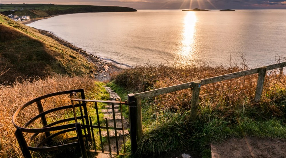 A view over a kissing gate with steps descending through grass to the shore, with the sea beyond and the sun low in the sky at Daron in Pwllheli, Gwynedd