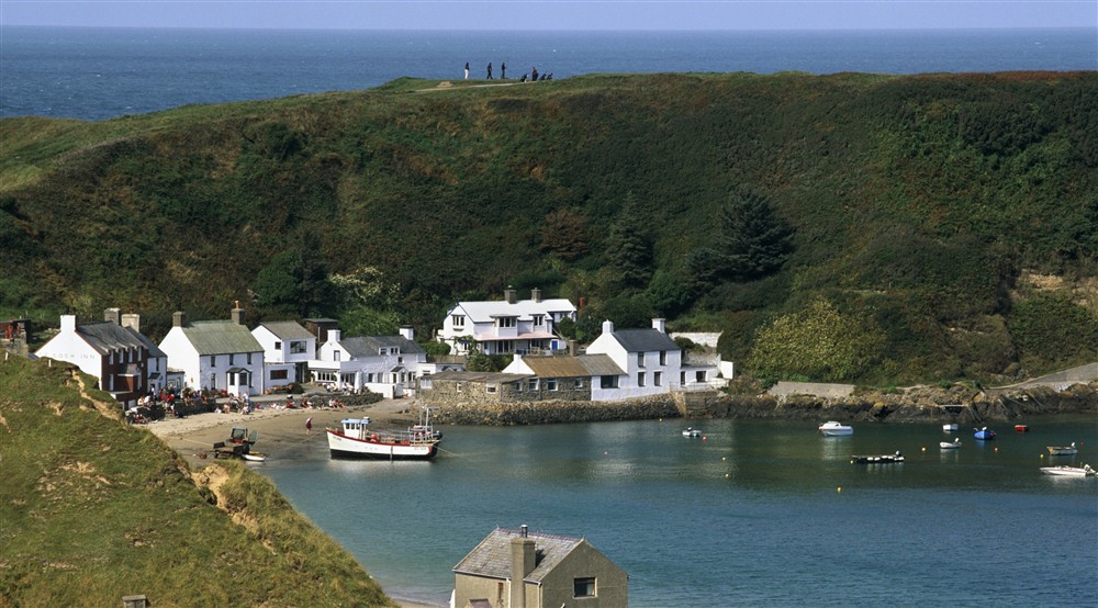A view towards Porthdinllaen, a small fishing village with a few white-painted cottages sitting beneath a grassy cliff on the Llyn Peninsula, Wales. Theres a few fishing boats in the water and people are walking on the cliff above the village. at Daron in