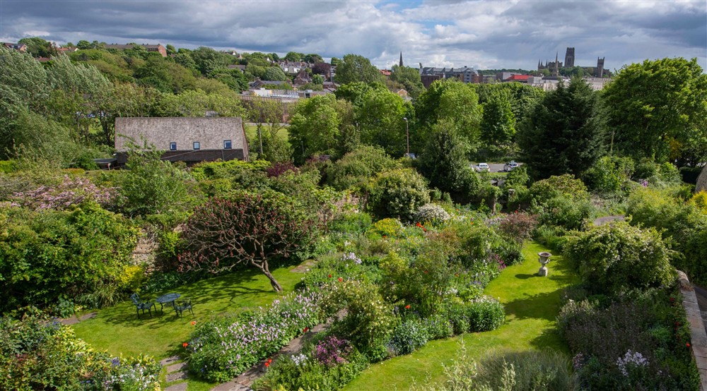 A wide aerial view over Crook Hall Gardens on a sunny spring day. Trees and shrubs are in full leaf, with colourful flowers in borders between green lawns. The roof of the café can be seen between the gardens and buildings on the edge of the city, with Du