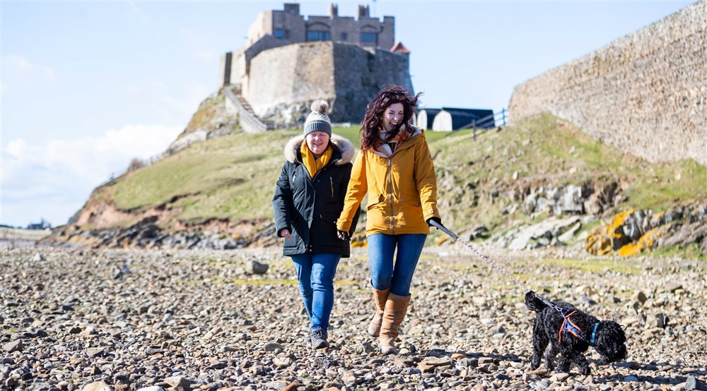 Two visitors walk along a pebbly beach with their black dog on a lead. In the background, a castle can be seen on top of the cliffs. at Glen House in Berwick-upon-tweed, Northumberland