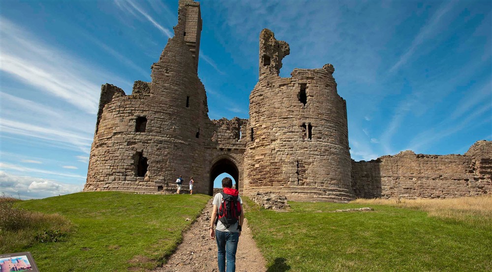 A visitor with their back to the camera walks along a path to the entrance of Dunstanburgh Castle at Lookout Cottage in Alnwick, Northumberland