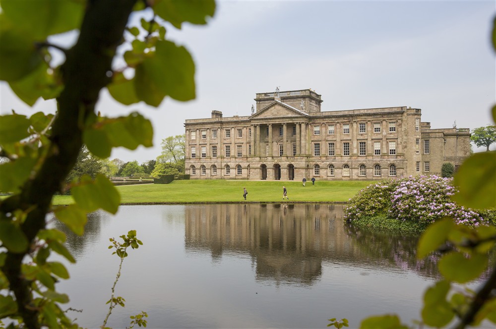 A view of the South Front of Lyme Park from across the lake with spring blooms framing the shot at Lyme West Lodge in Macclesfield, Cheshire