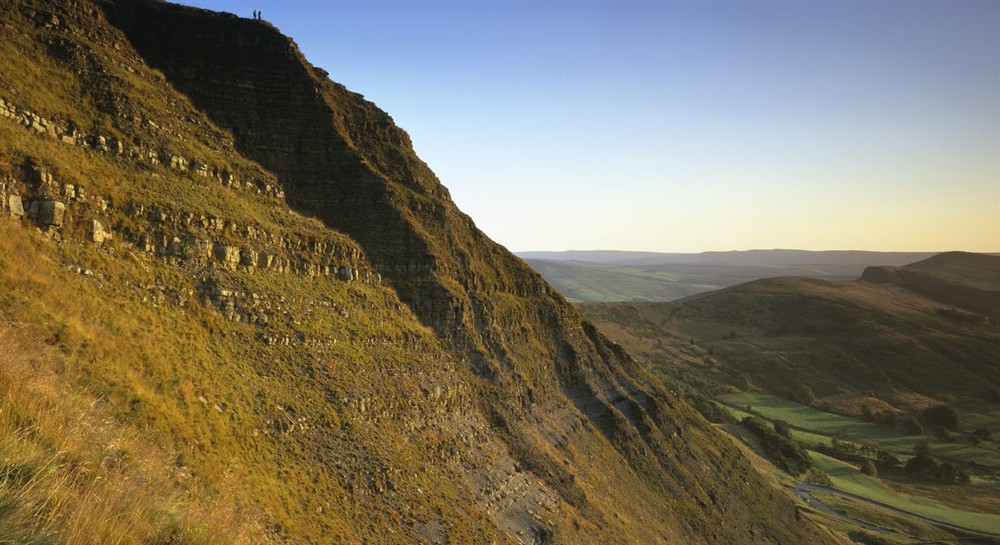 A steep sided mountain with a valley below in low early evening light. Two walkers in silhouette are just about discernible on the top of the mountain. at Mam Farm in Hope Valley, Derbyshire