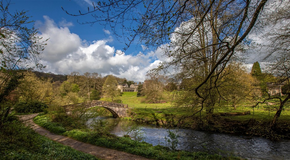 A view taken by the river through the trees across the stone bridge opening out into parkland, with views of Ilam Hall in the distance. at Mam Farm in Hope Valley, Derbyshire