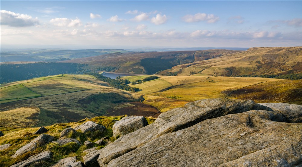 Sunny view with blue sky, from a rocky peak out over open moor and fields, with water in the distance at Mam Farm in Hope Valley, Derbyshire