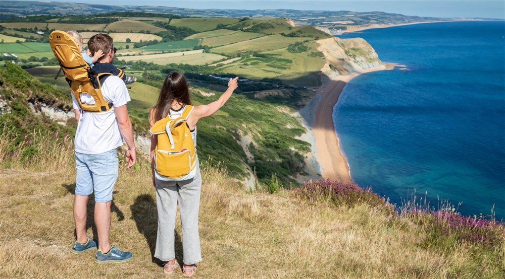 A woman and a man with a baby carrier looking out over the Jurassic Coast from Golden Cap in Dorset, on a bright summers day at Oak Cottage in Bridport, Dorset