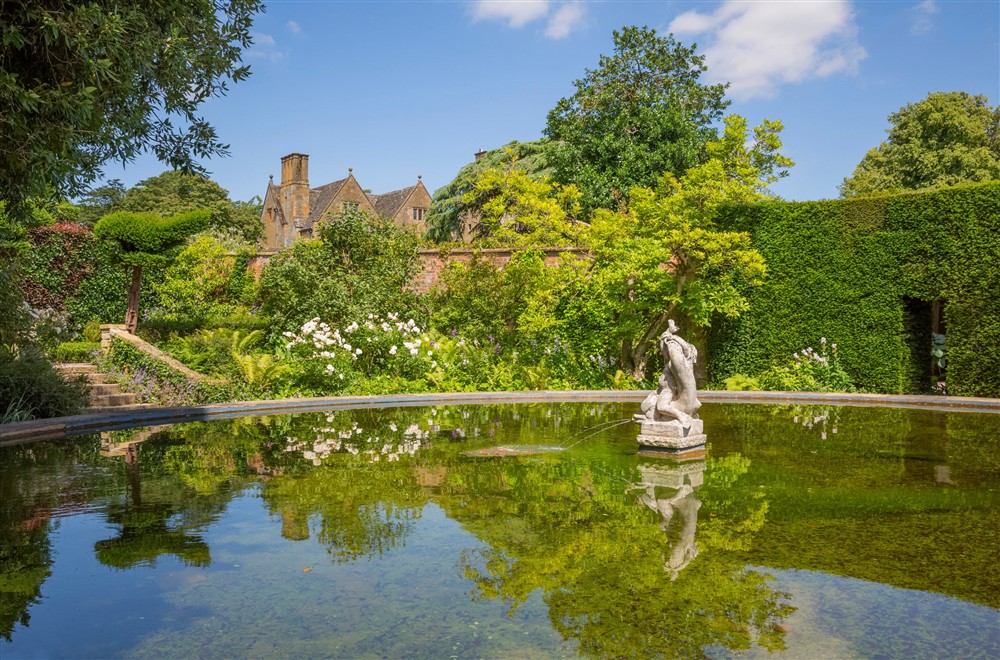A large circular pond with a stone fountain in the centre and a garden with trees and a manicured hedge behind, an old manor house in the background. at Spring Cottage in Broadway, Gloucestershire