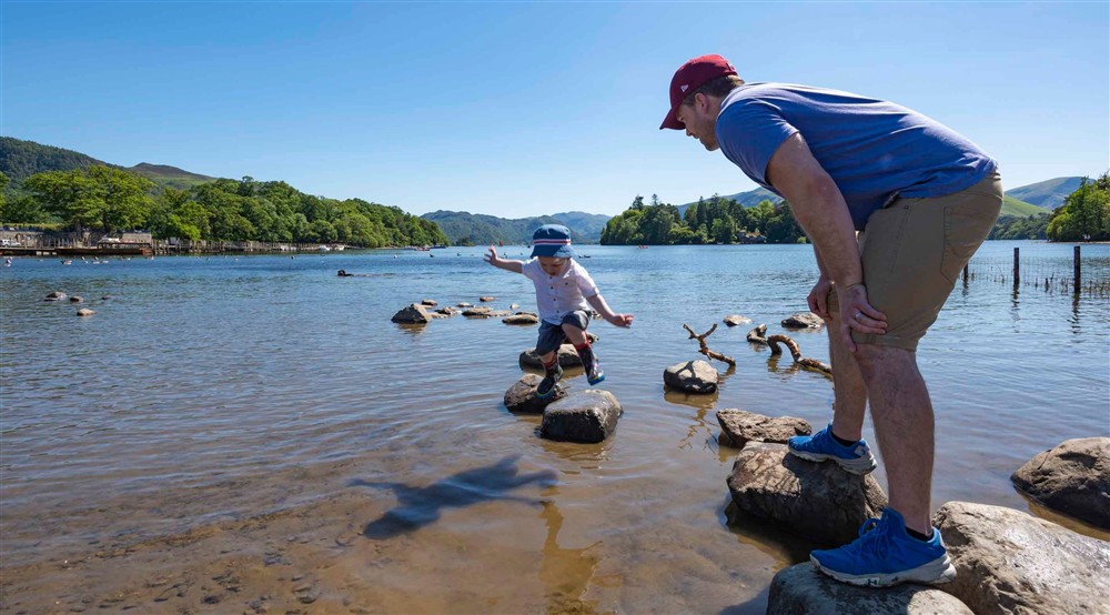 A father and young son standing on stones along the shore of Derwent Water at Watendlath Bothy in Keswick, Cumbria
