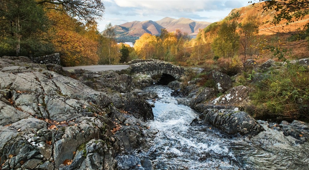 A stone bridge over a fast-flowing river flanked on either side with hard, grey stones and autumnal-leaved trees. Is the distance you can see warm evening light on dramatic-looking hills. at Watendlath Bothy in Keswick, Cumbria