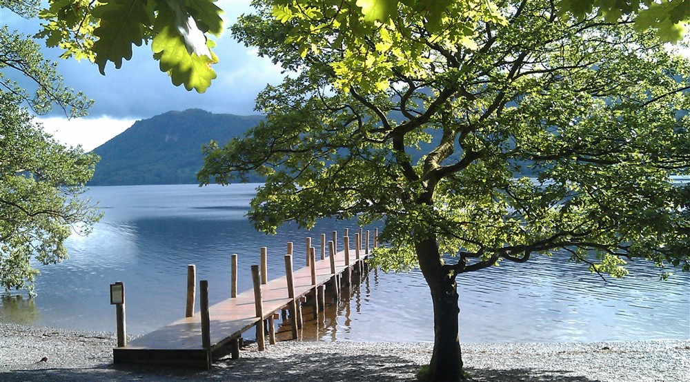 Sunny framed by green oak leaves, of a wooden jetty stretching out into sparkling Derwent Water at Watendlath Bothy in Keswick, Cumbria