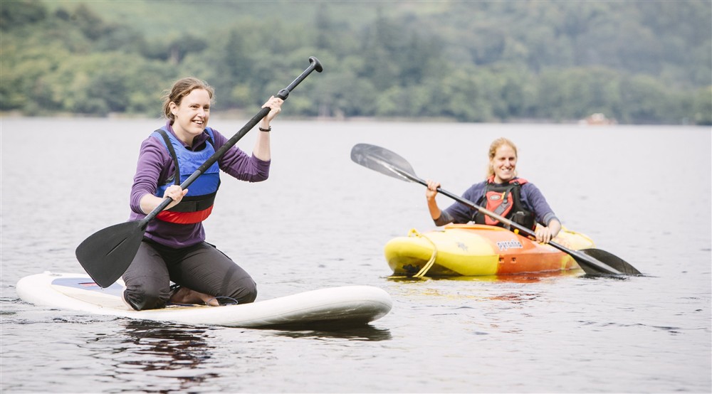 Two visitors enjoying watersports on Derwent Water: one paddleboarding and one kayaking, Lake District at Watendlath Bothy in Keswick, Cumbria