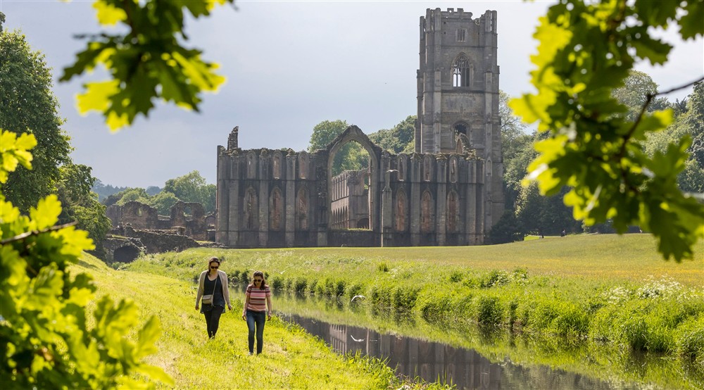 Two visitors walking through the grounds of Fountains Abbey and Studley Royal with the abbey ruins in the background at West Gate Lodge in Ripon, North Yorkshire