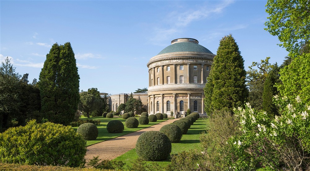 The rotunda sits in the middle of the picture with a path leading towards the bottom with round bushes lined up either side. there are tall green trees on the far left and right of the image with a blue sky overhead at Wicken Rose Cottage in Ely, Cambridg